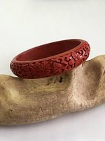 Side angled view of the red cinnabar carved bangle bracelet showing detailed floral vine carving and smooth interior surface placed on textured wood.