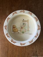 Top-down photo of ceramic baby bowl with brown bunny border and central painted design showing a duck and a bunny at a 'Paint Ball' stand