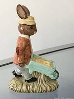 Side view of the Gardiner Bunnykins figurine showing the rabbit in orange jacket pushing a blue wheelbarrow on a grass-textured base.