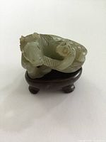 Front angled view of a small carved Chinese hardstone figure of a mythical or animal form placed on a round wooden stand with three legs.