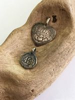 Two antique silver charms shown on wood background, one heart-shaped Claddagh pendant and one small round 'E' pendant