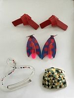 Top view of four vintage jewelry pieces: red clip earrings, purple/pink polka dot dangle earrings, clear acrylic bracelet with dots, pottery brooch.