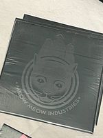 Photo showing close-up of embossed black cat face logo on a black card, reading 'Meow Meow Industries'.