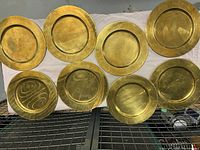 Eight round brass dinner chargers arranged on a surface, showing front side with reflective brass surfaces exhibiting wear and tarnish.