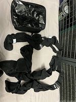 Top-down view of four black dog boots laid out with connecting straps, showing Velcro closures and fabric material, alongside a black carrying bag with clear front.