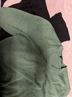 Green v-neck sweater folded on top of two black sweaters spread flat on a pink surface, showing fabric texture and color.