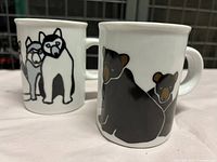 Front and side view of two white ceramic mugs with animal illustrations, one featuring black bears and the other huskies.