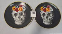 Two black decorative plates with a gold rim, each displaying a detailed skull illustration adorned with colorful flowers. Plates measure 10.5 inches.