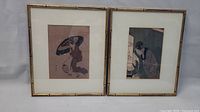 Two framed Japanese woodblock prints side by side against a grey background. Prints depict figures holding umbrellas, framed in faux bamboo gold frames with white mats.