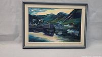 Framed village landscape painting depicting a waterfront village scene with houses, dock, and boats backed by hills, showing the full artwork and frame.