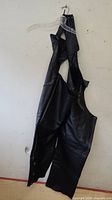 Black leather motorcycle chaps hanging on a clear hanger against a white wall. Shows front and overall shape.