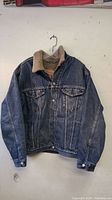 Front view of Levi Strauss jean jacket hanging, showing denim exterior and sherpa collar lining.