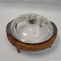 Side angle showing the wooden stand with three legs and the clear glass lid with metal rim and knob handle on top.