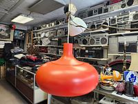 Orange pendant lamp with rounded, smooth, mid-century modern style shade in a matte finish, shown hanging with attached wiring inside a vintage radio and electronics store for context.