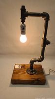 Full view of the lamp showing black metal plumbing pipes forming the arm with a light bulb hanging from a metallic silver socket above a polished wooden base.