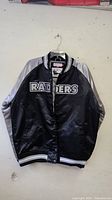 Front view of black and silver Oakland Raiders Mitchell & Ness NFL throwback jacket hanging on wall.