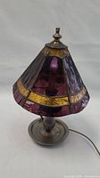 Front angle view showing stained glass shade with purple and amber panels and pewter base with round plate and handle.