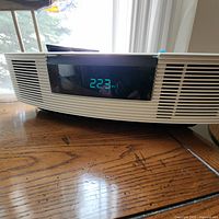 Front view of cream-colored Bose Wave Radio clock radio showing digital clock display at 2:23 PM. Horizontal grille vents on sides.