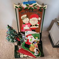 Photo showing assembled Christmas decor items including green lighted glass tree, Christmas stocking, wooden holiday sign with Santa and Mrs. Claus