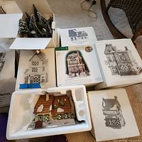 Overview of boxed and unpacked Dickens Village buildings and snow-covered pine trees in a box