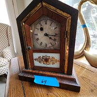 Front view of the steeple-style walnut mantle clock with Roman numeral face, gold trim, and decorative lower panel.
