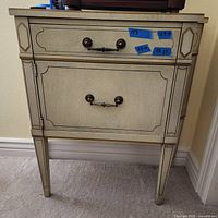 Front view of the cream-colored wooden end table with two drawers and decorative trim showing metal handles and tapered legs.