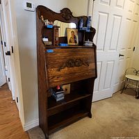 Full view of used wooden standing secretary desk positioned against a wall. The desk features a large mirror surrounded by carved wood, a drop front writing surface with decorative carvings, and two open shelves at the bottom.