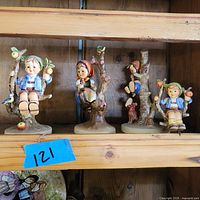 Four Hummel figurines displayed on wooden shelf, showing the full group for size and style comparison.