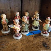 Collection of eight Hummel porcelain figurines posed on wooden surface with blue tape labeled '96'.
