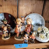 Photo of entire lot showing five Hummel figurines of children, two decorative plates with child and floral scenes, and porcelain rose floral arrangement