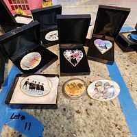 Six enamel pins displayed on a countertop, each in individual black boxes with cushions. Pins include a winter church scene, heart-shaped designs with couples, swans, and floral motifs.