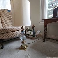 Full view of vintage side table lamp with white fabric lampshade placed on carpet near patterned armchair and wooden table.