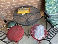 Photo showing black mesh metal round patio table with two folding chairs having red and green cushions.