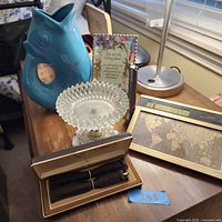 Photo of miscellaneous items on wooden side table including blue fish vase, framed religious quote, pens in box, glass plate, and Seiko world time clock