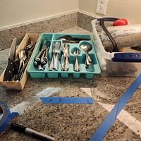 Wide view of silverware set with knives, spoons, forks, turquoise organizer, separate tray and electric meat carver on kitchen counter.