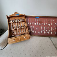Image showing wooden spoon rack holding eighteen silver plated souvenir spoons and two drawers below them; some spoons have detailed engravings on bowls and handles.