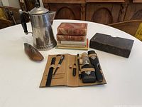 Overall view of the lot items on a white round table including metal coffee pot, books stacked, wooden shoe form, and toiletries travel case laid open with grooming tools.