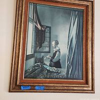 Photo of a framed wall painting featuring a woman in period dress standing by a window with draped curtains and a table with fruit, framed in a brown/gold layered frame.