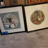 Two framed wall art prints leaning against a box on carpet. Left print shows children building snowman outside, right print shows couple kissing indoors.