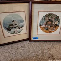 Two framed signed art prints side by side showing one winter church scene with carolers, one cozy interior with fireplace and family.