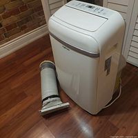 Full view of white EcoHouzng mobile air conditioner unit with vent hose laying beside it on wood floor.