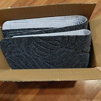 Photo shows a cardboard box filled with multiple gray textured no slip stair stick ons with an abstract or leaf pattern, sized to fit standard stairs.