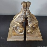 Photo showing front view of the two gold decorative book ends placed side by side, highlighting the symmetrical leaf and swirl design and the aged gold finish.