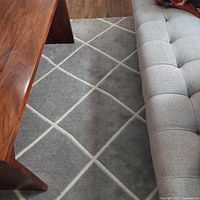 Close-up view of gray carpet with white diamond lattice pattern next to a gray couch and wooden coffee table, showing texture and design.