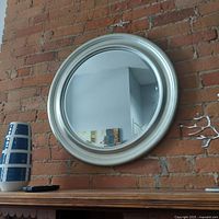 Round silver framed mirror hung on a brick wall, showing its reflective surface, wood base frame and dimensions approximately 27 by 27.5 inches.