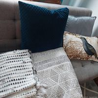Three decorative pillows arranged on a brown sofa including a blue velvet textured pillow, gray smooth pillow, and a bird printed pillow, alongside two throws draped over the sofa seat.