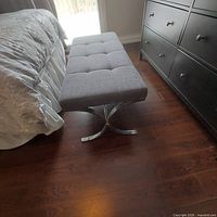 Side angle showing the bench's tufted upholstered grey fabric and polished metal curved legs beside a bed.