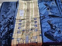 Three blue scarves laid out showing the Frank Lloyd Wright stained glass scarf in center, blue cut velvet scarf on left and large horse-themed silk square scarf on right.
