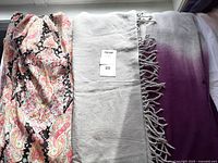 Photo showing three women's shawls laid out from left to right: a pink paisley light crepe-like shawl, a grey wool shawl with beads, and a purple and grey cashmere mix shawl with fringe and buttons.