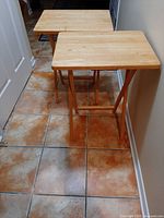 Two wooden TV trays standing side by side, showing overall shape and wood finish on tiled floor.